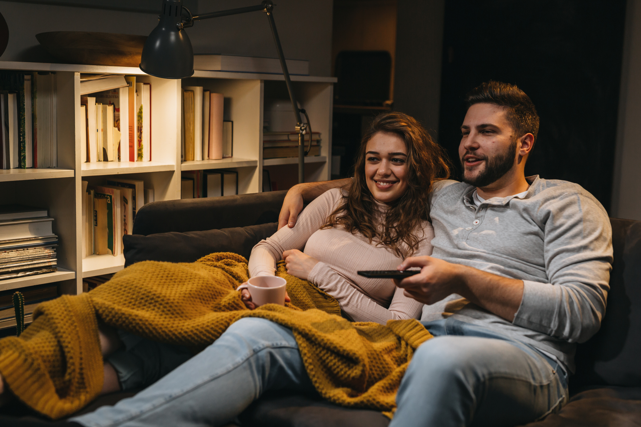 A man and woman sit out a couch underneath a yellow blanket. The woman is holding a coffee mug and the man is holding a TV remote. They are both looking away from the camera, presumably towards a television screen.