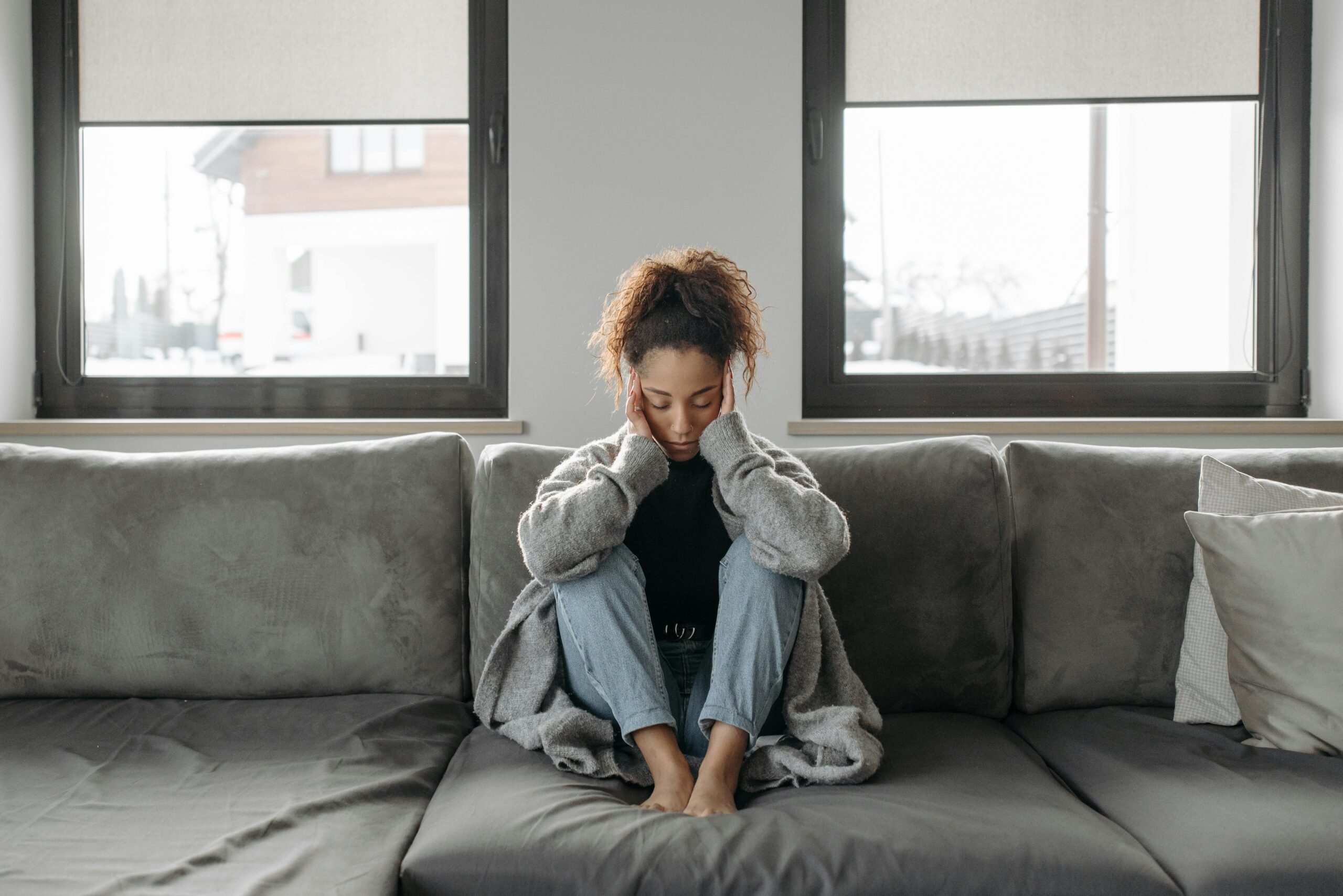 A young woman with medium-dark skin and curly brown hair sits on a couch. She is wearing a large sweater and holding her head as if she has a headache.