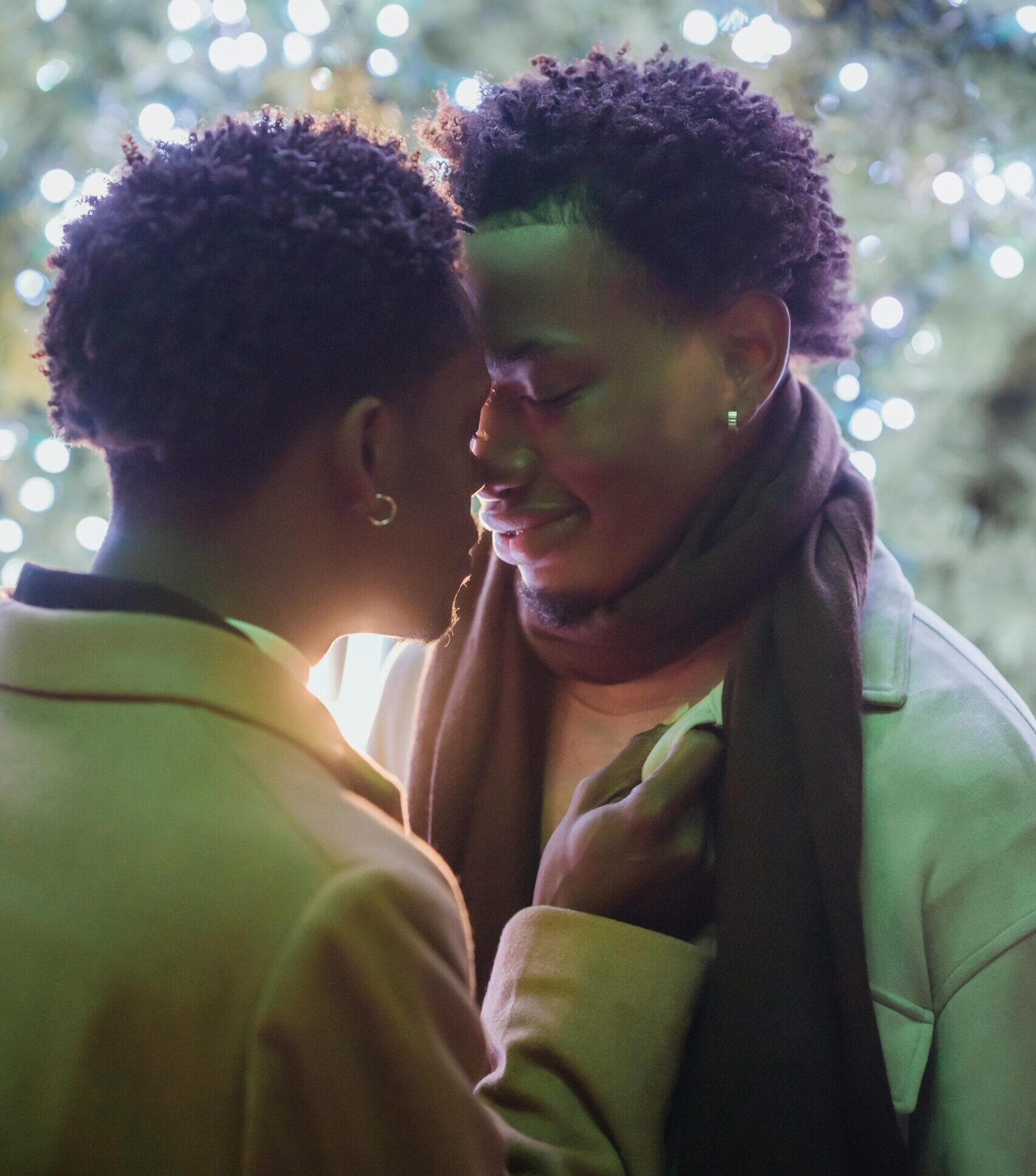 Two Black men who each have short, kinky hair embrace in front of a tree decorated with lights.