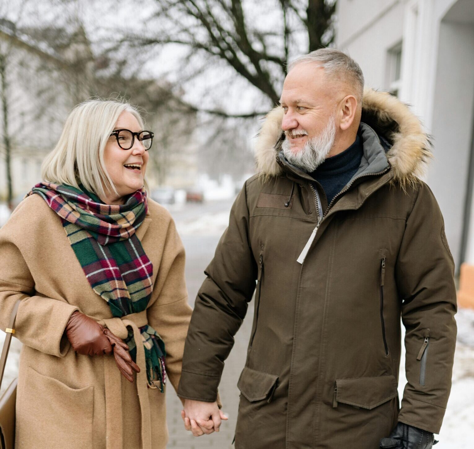 An older white woman with straight blonde hair and glasses holds hands with an older white man with a beard. They are standing on a snowy street and smiling at one another.