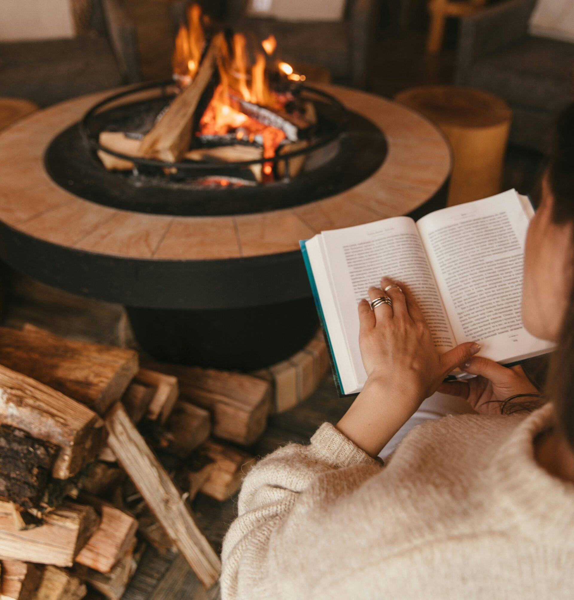 A white woman wearing a cream-colored sweater sits in a chair in front of a fireplace and reads a book.